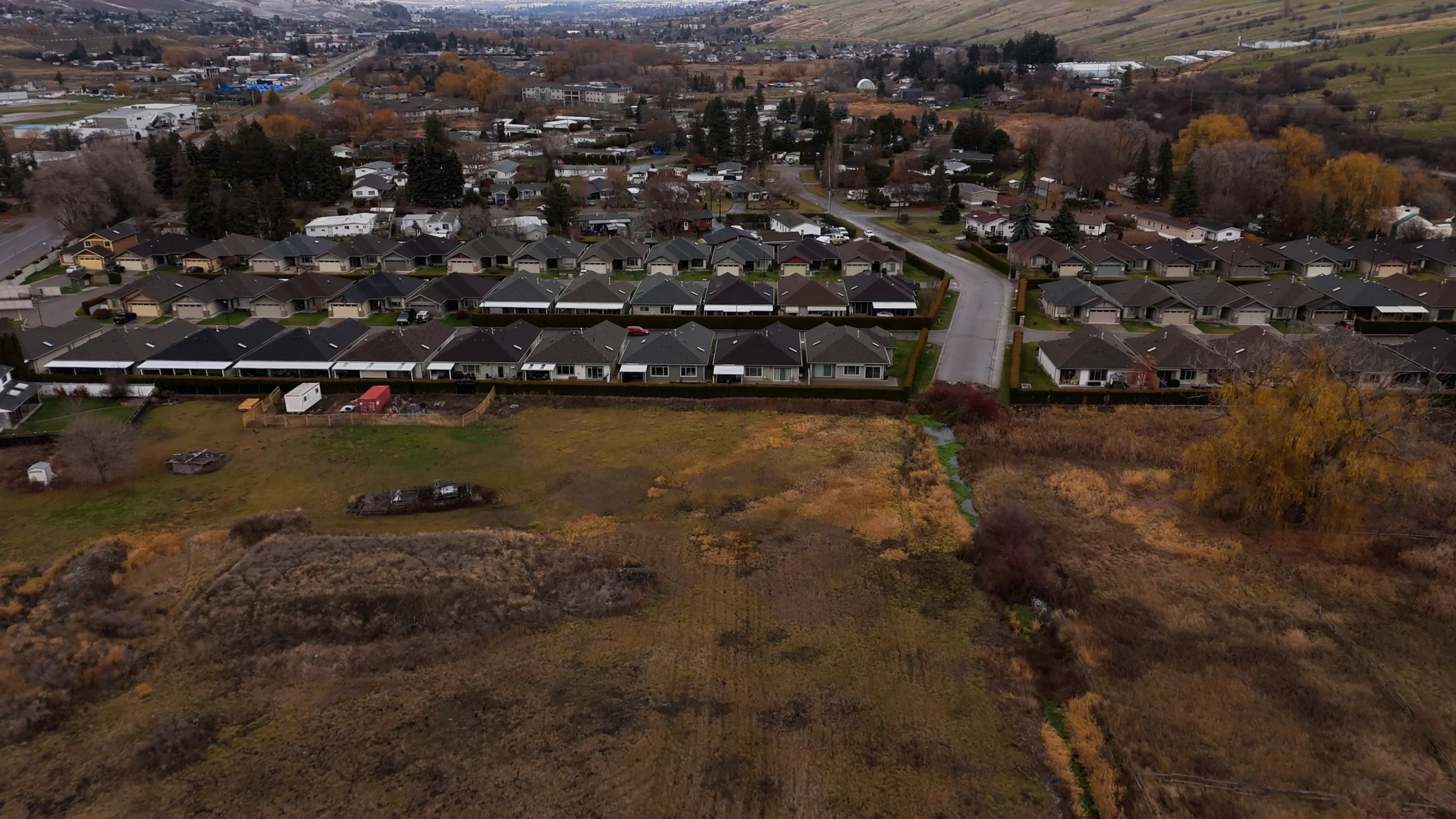 Aerial view of suburban homes bordering an open field and creek, with hills and neighbourhoods in the distance.