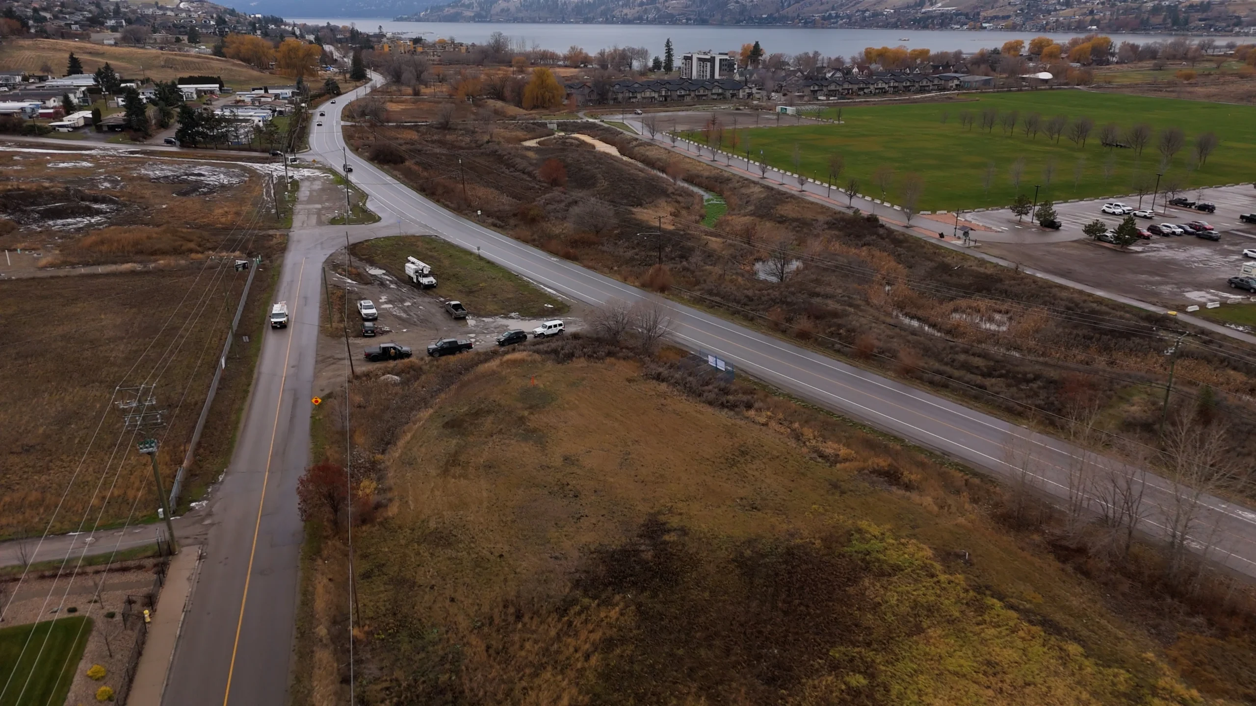 Aerial view of open land and roads near a park, with Okanagan Lake and hills in the distance.