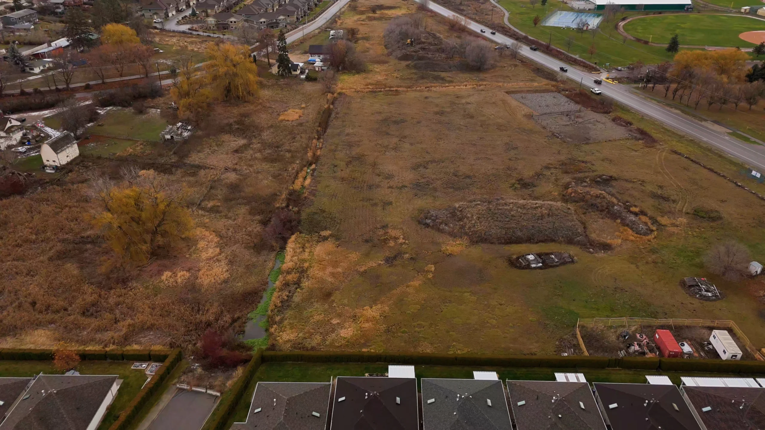 Aerial view of a vacant field with a creek, bordered by homes and a road near a sports complex.