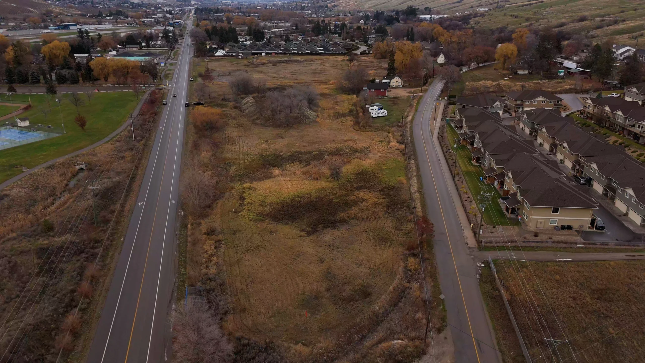 Aerial view of vacant land between two roads, with townhomes, park fields, and hills in the background.