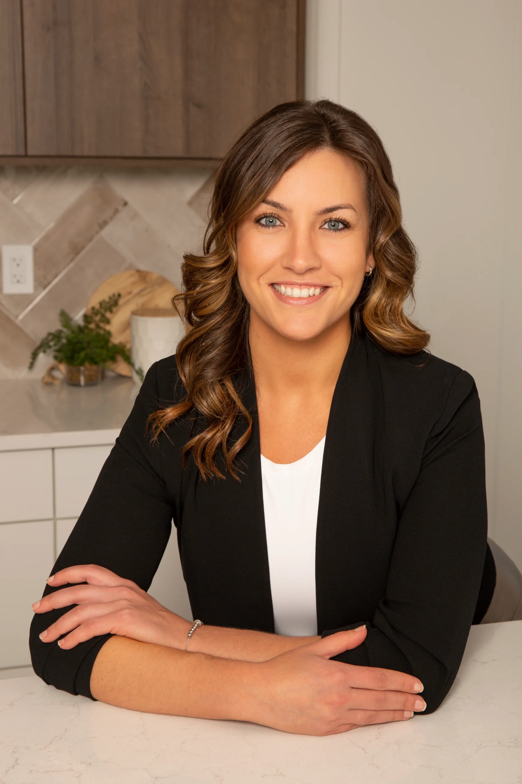Professional headshot of a woman in a black blazer, smiling with arms crossed in a modern office setting.