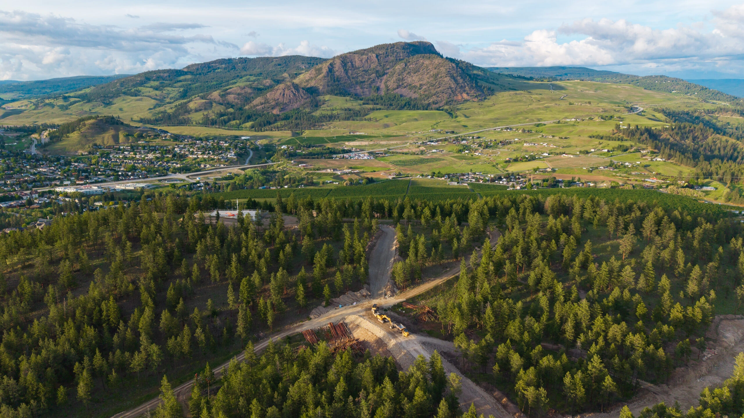 Aerial view of forested hillside construction with access roads, overlooking Okanagan Valley farms and a mountain ridge.