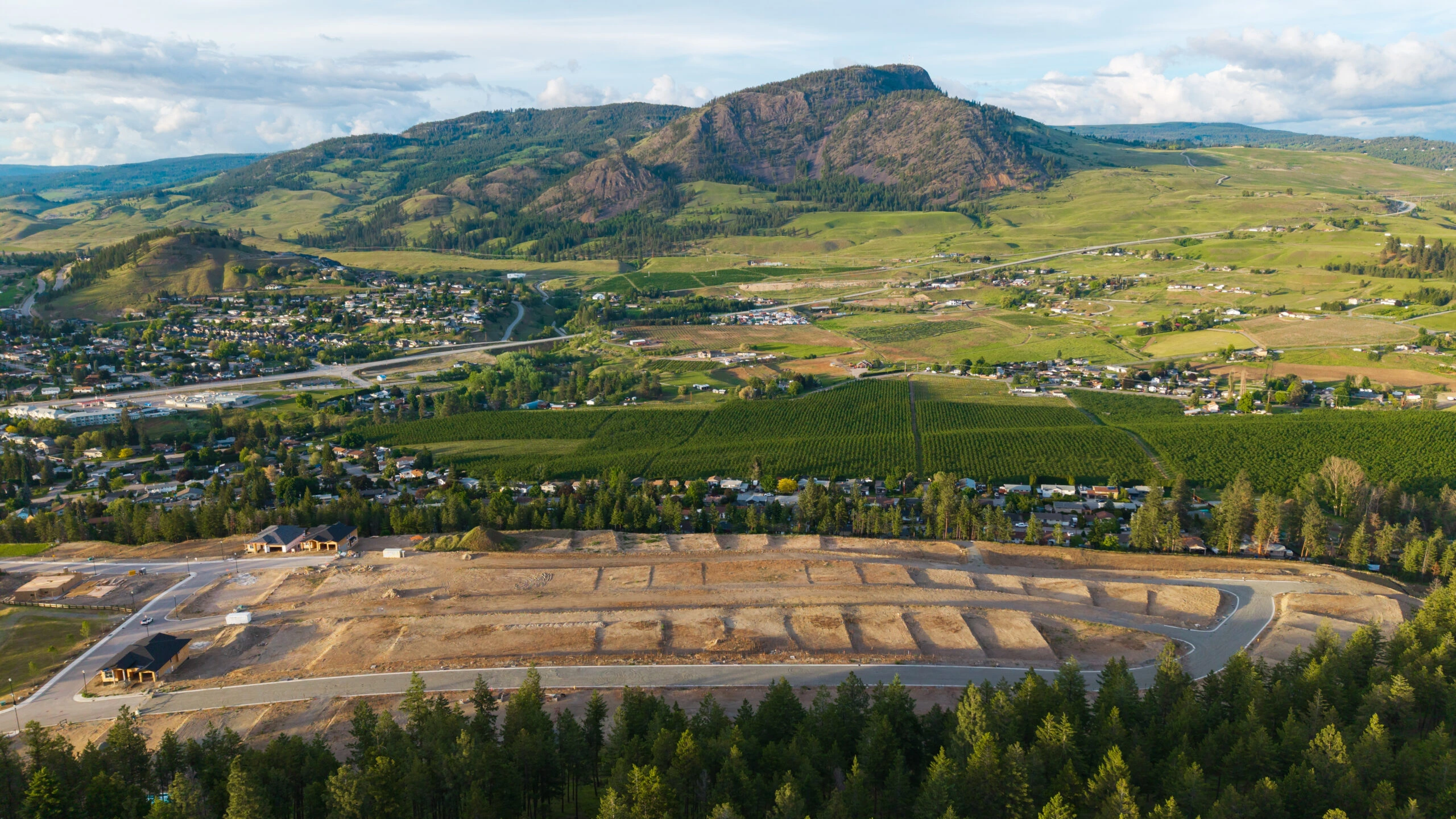 Aerial view of a graded subdivision site above orchards and neighbourhoods, with Okanagan Valley and mountains beyond.
