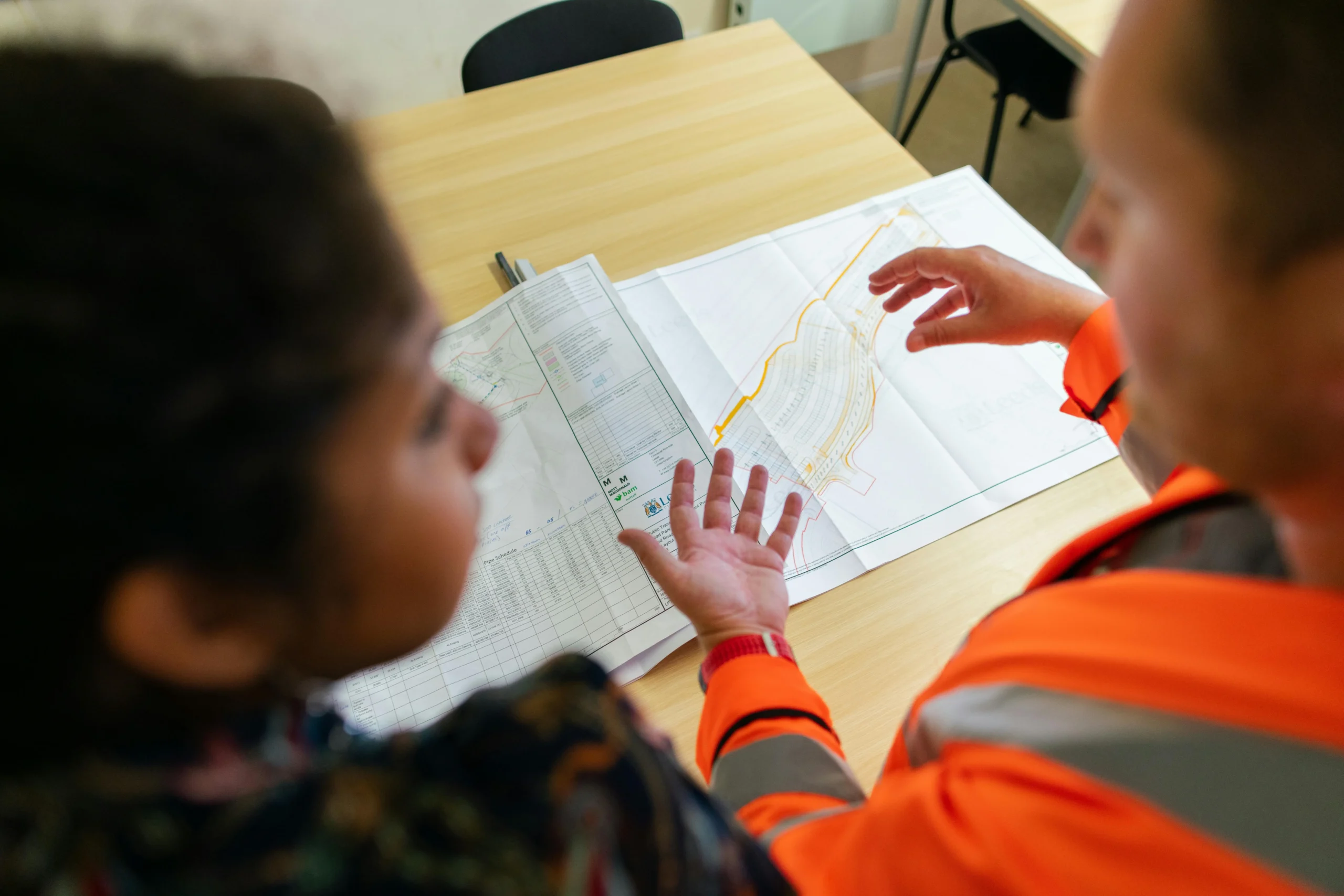 Close-up of an engineer in high-vis orange and a partner reviewing detailed development plans on a table, gesturing to site layouts.