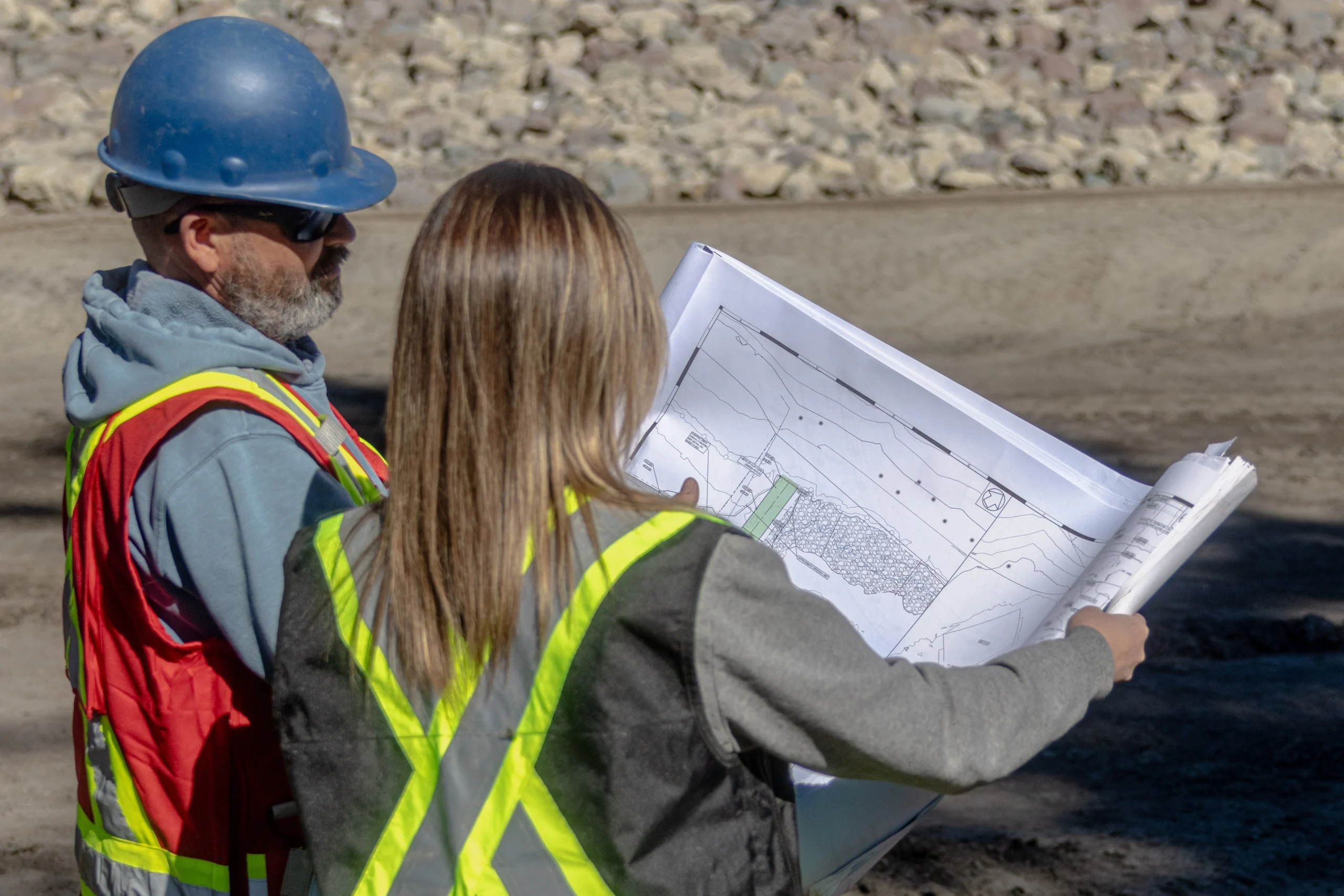 Two professionals in safety vests and a hard hat review architectural blueprints on a sunny outdoor construction site.